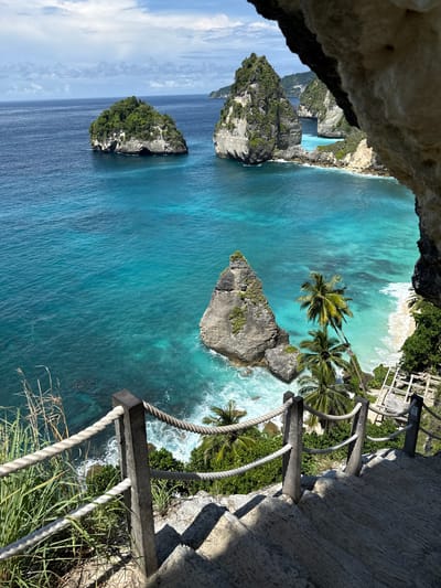 The image depicts the landscape of Nusa Penida, a small island southeast of Bali, Indonesia, specifically in the city of Pejukutan. The view is from a high vantage point, looking down over the coast. Turquoise water dominates the scene, contrasting with rocky islands covered in greenery. A white sand beach is visible in the distance. The image is taken from what seems like a cave or natural overhang, with a rocky wall on one side, and a man-made rope fence is present on the other. The bright sunlight casts long shadows.
