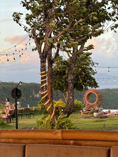 The image depicts an outdoor leisure area at dusk in Sakti, Indonesia, overlooking a bay or sea surrounded by forested cliffs. In the immediate foreground, a bamboo and light-brown cushioned barrier, possibly part of a seating arrangement, extends horizontally across the frame.

The midground features a manicured green lawn dotted with various elements. Two prominent trees with visible trunks and green foliage stand near the center, one wrapped with a spiral of illuminated warm-toned string lights. Overhead, multiple strands of string lights with exposed bulbs crisscross the scene.

On the left side of the lawn, a black pole with a circular sign stands erect. Behind it, two individuals are seated at a dark table with chairs. One person, with light-colored hair, is seated facing away from the viewer, looking at a phone. Another individual, wearing a dark shirt and a bandana, stands nearby, holding an object resembling a drink or device.

Towards the right midground, a large, circular decorative structure made of woven rattan or natural fibers, resembling a stylized sun, is positioned on a wooden base. Adjacent to it, two individuals are reclining on low-level lounge furniture or mats on the grass. One person, wearing a light-colored top, holds a bottle. A white planter pot is situated on the grass nearby.

The background consists of dense green foliage covering steep cliffs or hills. Beyond these landforms, a calm body of water is visible. The sky above displays soft cloud formations with hints of pink and orange, indicative of either sunset or sunrise.