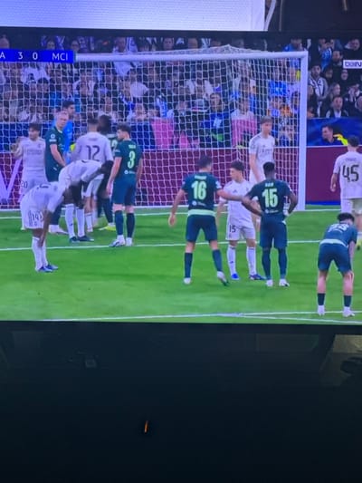 The image presents a photograph of a television screen displaying a soccer match, captured within Vitry-sur-Seine, France. The upper left corner of the screen shows a scoreboard indicating "A 3 0 MCI." The scene centers on a section of a green grass soccer pitch, featuring a white goal net and red-brown advertising boards at its base.
Several male soccer players are visible on the field. One group wears white jerseys, sponsored by "Emirates Fly Better," with discernible numbers 20, 14, 12, 15, and 45. Player 14, in a white jersey, is bent at the waist, resting his hands on his knees. Player 45 in white has "THIAGO" printed above his number. Another group wears dark green/teal jerseys, with visible numbers 3, 16, and 15. Player 3 has "TREN" or "RUBEN" (partially obscured) above his number, and Player 15 has "BEN" above his number. Players 16 (green) and 15 (green) are standing with their backs partially to the camera, while player 15 (white) is positioned in front of player 16 (green).
In the background, behind the goal net, stadium seating filled with blurred spectators is visible. A portion of a "BEIN" logo is discernible on an advertising banner in the upper right background. The lower third of the overall image is dark, depicting the television screen's bezel or the surrounding room.