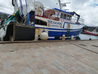 The image depicts a fishing trawler named "HEGOAK" docked in the port of Cherbourg-en-Cotentin, France. The vessel is painted blue and white and is equipped with various fishing gear, including nets and buoys. The dock is paved with granite cobblestones. In the background, another red and white fishing boat is visible, also docked. The sky is overcast with clouds.