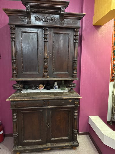 The image depicts an antique, dark wood hutch against a vibrant magenta textured wall. The hutch is ornately carved with decorative elements, including floral motifs and turned columns. It features two large cabinet doors on the upper section and two smaller doors on the lower section, separated by a narrow drawer. On a shelf above the lower doors, several small decorative items are displayed, including a teapot, a black vase, and a small floral patterned vase. To the right of the hutch, a portion of a colorful tapestry or rug is visible hanging on the wall, and a yellow architectural element extends from the upper right corner of the frame. A portion of a metal railing or furniture is visible on the far right. The floor appears to be carpeted. The overall scene suggests an indoor setting, possibly a room in a museum or a home decorated with antique furnishings. The specific location in Fort-Şevtşenko, Kazakhstan, is not visually ascertainable from the image itself.