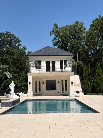 The image depicts a modern luxury home and its outdoor amenities. The scene includes a sleek, multi-story house with light-colored stone facade, dark grey roof, and numerous large windows. A swimming pool with turquoise water is adjacent to the house, its surface rippling. The pool area is paved with light-colored stone tiles.
Surrounding the house and pool are lush green trees, indicating a possibly wooded or well-landscaped environment. Several white spherical planters with greenery are strategically placed around the property. A white sculpture, possibly of a reclining figure, is visible in the foreground, adding an artistic touch to the outdoor space.
The time of day appears to be daytime, with bright sunlight casting shadows and illuminating the scene, suggesting fair weather. The overall impression is one of opulence and tranquility. No visible text is present in the image. The location context provided is Potomac, USA.
