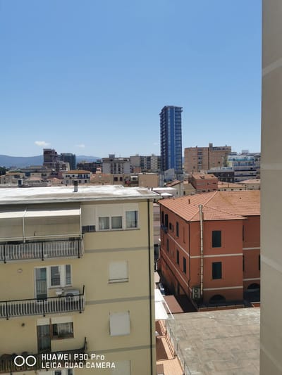 This is a daytime, eye-level, outdoor shot of a cityscape in Follonica, Italy, featuring multiple buildings under a clear blue sky. In the foreground, on the left, is a light yellow building with white balconies and white-framed windows, some equipped with white rolling shutters. An air conditioning unit is visible on one of the balconies. Adjacent to it, on the right, is a reddish-orange building with multiple windows, some with dark shutters. The top of this building features a terracotta tiled roof with a metal chimney. In the middle ground, a cluster of various residential and commercial buildings of different heights and colors are visible, including a tall blue skyscraper in the background. A mountainous landscape is faintly discernible in the distance. The ground level beneath the buildings is not clearly visible. The lower right corner of the image displays the watermark "HUAWEI P30 Pro LEICA QUAD CAMERA". There are no people or animals present in the scene.