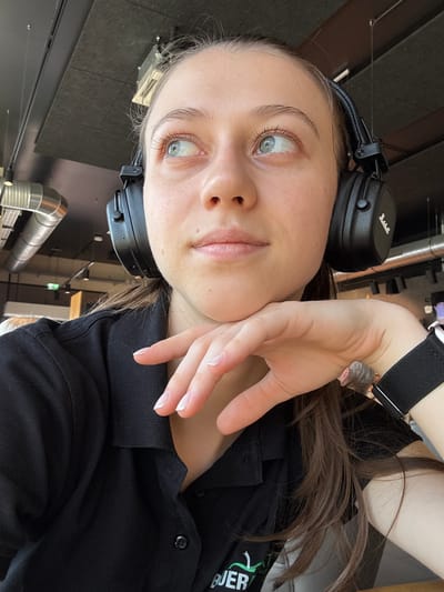 The image is a close-up, vertically oriented selfie of a young woman with light skin and long, brown hair. She is wearing black over-ear headphones, which are visible on her head and partially obscuring her hair. Her right hand is resting on her cheek, with her fingers extended and gently touching her face. She has light blue eyes and her lips are slightly parted. She is wearing a black collared shirt. A small portion of a black wristband with a silver buckle is visible on her left wrist. The background is blurred but appears to be an indoor setting with industrial elements, including metal piping and a dark, tiled wall. The lighting suggests it is daytime, possibly indoors with artificial lighting. There is a subtle sheen on her skin, indicating either natural light or a dewy makeup effect. The overall impression is a casual, personal moment captured by the subject. There is some partially visible white text on the right side of her shirt, but it is not legible. The location context is Elsenfeld, Germany.