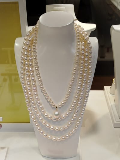 A layered pearl necklace is displayed on a white mannequin bust in a store. The necklace consists of multiple strands of pearls, ranging in size from small to large, with the largest pearl suspended as a pendant. The pearls have a lustrous, iridescent sheen. The mannequin bust is positioned in front of a yellow background and a dark background. To the right of the mannequin, another white display stand is partially visible with a gold ring on it. The scene appears to be a retail display, likely in a jewelry store. There are no people visible in the image. The location is specified as Marki, Poland, but this information cannot be visually confirmed from the image itself.