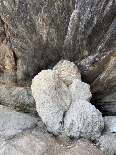 The image shows a natural rock formation in Dom Otdykha Zolotoy Plyazh, Russia. The formation is composed of large, rough-textured rocks. In the foreground, a cluster of pale, coarse-grained rocks is prominent. Behind these, darker, more layered rocks form a backdrop. The overall scene is outdoors, with visible elements of a rocky, natural landscape. There are no people or man-made objects visible in the image. The lighting suggests it is daytime.