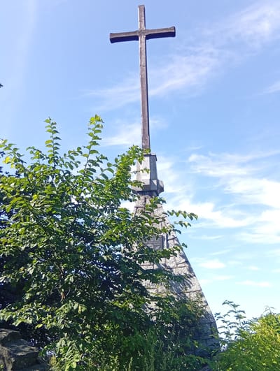 A wooden cross stands atop a stone structure against a bright blue sky with scattered white clouds. The cross is dark brown and appears weathered, with a vertical post and a shorter horizontal beam. The stone structure is triangular in shape, made of rough, light-colored stones and appears to be part of a monument. Lush green foliage, likely trees and bushes, obscures the lower portion of the stone structure and extends from the left side of the frame, reaching towards the cross. The top of a portion of the cross is visible in the top center of the image. The sky is a clear blue with thin, wispy clouds.