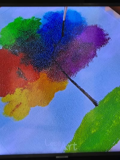 The scene, located in Zangiata, Uzbekistan, displays a close-up photograph of a digital screen, identifiable by a black bezel and a "SAMSUNG" logo centered on its bottom edge. The screen exhibits an artwork in progress, resembling an oil or acrylic painting on a light blue textured canvas background. A central cluster of distinct, thickly applied paint blobs is visible, featuring hues of red (lower left), yellow (mid-left/bottom-center), green (upper left and lower right), dark blue (upper center), and purple (mid-right). A paintbrush, with a metallic ferrule and dark bristles, is visible entering the frame from the top, angled downwards towards the center of the paint cluster. It is actively applying black paint, forming thin, radiating strokes from the central mass of colors, with one prominent stroke extending diagonally towards the lower right corner. Faint white text, possibly "Leege Art," is subtly present in the lower central area of the displayed artwork. No people are visible.
