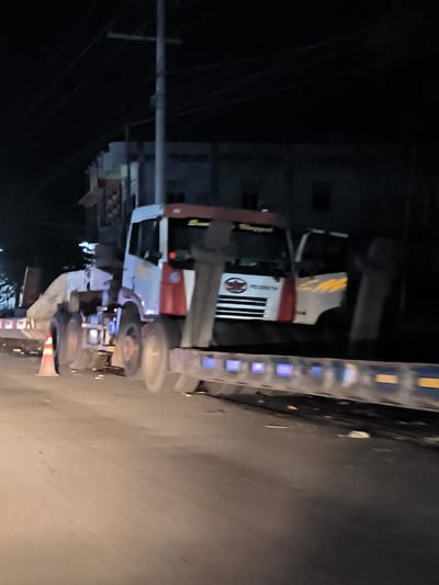 The image captures a dark, low-light scene on a road at night, likely in an urban or semi-urban area within Meuse, Indonesia. The central subject is a large white and red truck, possibly a heavy-haul truck or a prime mover, viewed from its front-left side. It appears to be transporting a substantial, irregularly shaped, light-colored load, possibly industrial equipment or a large component, on a multi-axle flatbed trailer with reflective blue and yellow squares along its side.
The truck's wheels show some motion blur, suggesting it is either moving slowly or the photo was taken with camera shake in low light. An orange traffic cone stands on the road near the truck's front left wheels, indicating a temporary stop or a designated area. In the background, dark, indistinct buildings are visible against the night sky, partially obscured by a tall utility pole and numerous overhead wires. Another smaller white vehicle, possibly a pickup truck, is partially visible behind the main truck.
Notable details include the very dark conditions, with artificial light sources illuminating only parts of the road and vehicles. On the front grille of the main truck, a circular logo is present, with some barely discernible text below it, possibly "FD 230 TF." Above the windshield, some blurry dark text can be seen on the white cab. The overall impression is one of a nighttime heavy transport operation or a roadside stop.