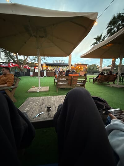 The scene depicts an outdoor dining or social area in Havat Shalem, Israel, observed from a low perspective under an overcast sky, indicating late afternoon or early evening.
In the immediate foreground, the legs of two individuals are visible. The person on the left wears dark-colored pants, and the person on the right wears light blue clothing, holding a smartphone in their lap. A slatted wooden table is positioned between them, holding a small dark-colored jar and a light-colored utensil, possibly a spoon or small knife.
The midground consists of a large expanse of artificial green turf. Scattered across this area are numerous light-colored patio umbrellas with central poles, along with various wooden benches and chairs. To the left, a male individual in an orange hoodie is seated at a wooden table, facing left and exhibiting a smile. Further back, several other individuals are seated at additional wooden tables and benches, some featuring orange cushions, engaging in social interaction.
The background reveals a row of casual structures or stalls. A prominent sign displaying the word "VANV" is visible on a central structure, likely a food or beverage service point. Other stalls exhibit features such as red umbrellas or awnings. String lights with exposed bulbs are strung overhead across the area. Palm trees are visible on the right side against the sky, while other trees and foliage are present on the far left. The sky is predominantly grey with hints of blue.