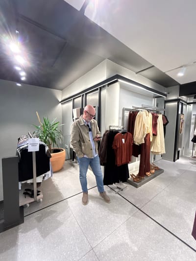 An interior view of a modern clothing retail store in Chaves, Portugal, features a bald male subject with glasses standing in the center-left. He wears a light blue collared shirt, a beige jacket, light blue jeans, and light brown suede shoes, actively viewing a smartphone held in both hands. To his left, a mobile clothing rack displays various dark garments and a white sign labeled "TAMANHOS ÚNICOS" (Unique Sizes), with black high-heeled shoes on a lower platform. Behind this rack, a large green plant sits in a terracotta pot. To the man's right, a stationary clothing rack presents an array of earth-toned garments, including rust-colored tops, beige shirts, and rust-colored trousers. Three pairs of light brown high-heeled shoes are neatly arranged on the floor in front of this rack. The store environment is characterized by light gray walls, a dark gray ceiling with recessed spotlights producing lens flares, and linear LED lighting strips along wall fixtures. The floor is composed of light gray speckled tiles with dark grout lines. A small security camera is visible on the upper right wall.