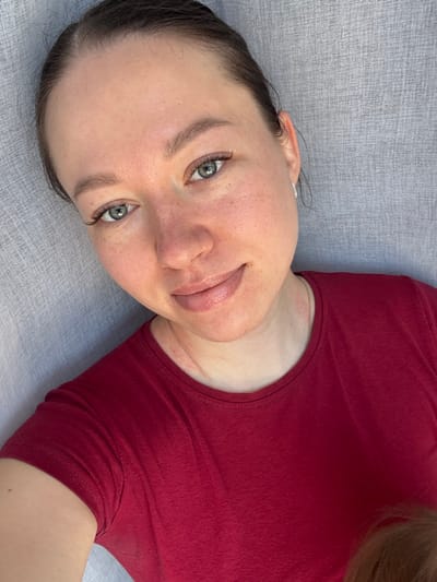 An adult female is depicted in a close-up, selfie-style indoor portrait, positioned diagonally, looking directly at the viewer with a slight, closed-lip smile. She has light skin with subtle freckles across her nose and cheeks, green-blue eyes enhanced with what appear to be eyelash extensions, and light brown eyebrows. Her dark hair is pulled back from her face. A small, silver-colored hoop earring is visible in her right ear. She wears a plain, short-sleeved, maroon crew-neck t-shirt. A faint gold-colored chain is visible beneath her collar. Minor red skin irritations are present on her neck and upper chest. In the lower right corner, a partial view of light-colored hair, consistent with that of a child, is discernible. The background is a uniform, light gray textured surface, possibly fabric. The scene is an unidentified indoor location; no visual information indicates the city of Shul'gino, Russia.