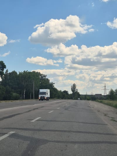 A white truck with a box trailer is driving on a paved road in Pidhaitsi, Ukraine. The road has white lane markings and appears to be a two-lane highway. To the left of the road is a dense line of green trees, including some birch trees. To the right of the road, there is a grassy shoulder and more trees in the distance. In the far background, there are power lines and what appear to be industrial structures. The sky is blue with scattered white, fluffy clouds. The lighting suggests it is daytime, likely late afternoon due to the warm tone of the light.
