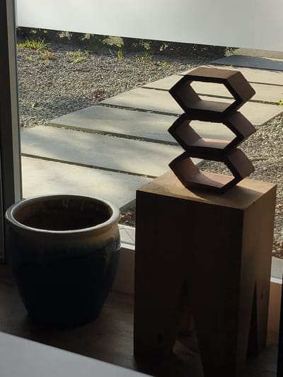 The scene depicts an interior view looking out through a window onto an exterior landscaped area in Riccione, Italy. In the immediate foreground, a dark-glazed ceramic planter with a lighter, stained rim sits on a dark wooden surface to the left. To its right, a tall, dark wooden pedestal with a block-like form and visible angular cut-outs near its base supports an abstract sculpture. The sculpture consists of three stacked, dark brown, hollow hexagonal elements. The topmost two hexagons are fully visible, while the bottom one is partially obscured by the pedestal.
Through the clear window, an exterior ground area is visible. This area is covered predominantly with small, light-colored gravel. A path of several flat, rectangular concrete pavers is laid across the gravel. Sparse green vegetation, primarily small grasses, grows within the gravel. In the background, a low, light-colored horizontal structure or wall is present, behind which dense green foliage can be seen. Strong, natural light illuminates the exterior, casting distinct shadows, particularly from the concrete pavers, while the interior objects are largely in shadow. No people or active interactions are present in the image.