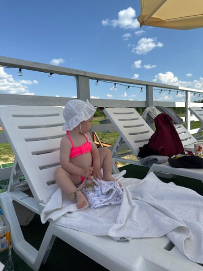 A baby wearing a pink bikini and a white sun hat lies on a white plastic lounge chair covered with a white towel. The baby is on their back, with legs bent and feet facing upwards. A white cloth with a floral pattern is tucked under the baby's legs. A pacifier with a beaded string is attached to the baby's hand. In the background, there are other empty lounge chairs, some with clothing draped over them, and a wooden structure with string lights. The scene appears to be outdoors, possibly at a resort or recreational area, with a clear blue sky and white clouds visible above. The lighting suggests it is daytime. There is no visible text in the image.