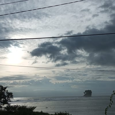 A wide shot captures a hazy, overcast day at the coast in Keudeunga, Indonesia. The sky is dominated by a mix of dense, dark clouds and lighter, wispy formations. A bright, diffused sun is visible on the left, partially obscured by clouds. In the distance, a small, rocky island topped with dense foliage stands in the ocean. The sea is calm with gentle waves. In the foreground, the tops of trees and foliage are visible on the left and right sides of the frame, with a few dark lines, likely power lines, stretching horizontally across the sky. The overall atmosphere is subdued and slightly muted due to the cloudy weather.