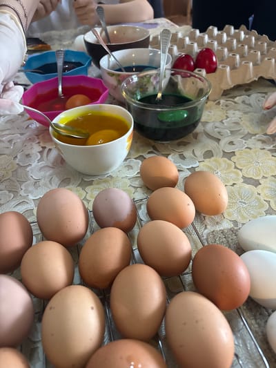 The image shows a table with eggs being dyed, likely for Easter. There are bowls with different colored dyes (yellow, green, blue, pink) and spoons. Raw eggs are nearby, some on a cooling rack. An egg carton with dyed red eggs is also visible. The table is covered with a floral patterned cloth. The location is not discernible from the image and thus cannot be identified as Makariopolsko, Bulgaria.