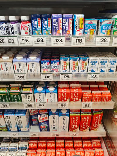 The image shows a retail display of dairy products, predominantly milk and yogurt, on shelves in a brightly lit store. The products are mostly packaged in cartons and bottles, with Japanese text and branding visible. There are no people in the image, and the focus is solely on the merchandise. The shelves are well-stocked and organized, suggesting a commercial setting. The overall impression is that of a typical grocery store or convenience store aisle. The specific location of Sapporo, Japan, is provided as context, but there are no visual cues in the image itself to confirm this geographical detail. The products range in size and type, indicating a variety of choices for consumers. The lighting is even and reflects off the packaging, highlighting the product labels.