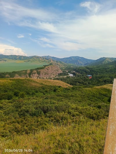 The image shows a vast, green landscape with rolling hills and a valley. In the foreground, there is a grassy hillside with tall dry grass and lush green bushes. Further in the distance, a rocky outcrop juts out from a greener hillside. Beyond this, a valley opens up, filled with dense green trees. A river winds through the valley, with a few buildings visible on its banks, including a blue-roofed building and a red-roofed building. In the far distance, a series of hazy, green mountains stretches across the horizon under a blue sky with scattered white clouds.
The scene suggests a natural, rural setting. The time of day appears to be afternoon, indicated by the brightness of the sky and the shadows cast. The weather seems pleasant, with a clear blue sky and only a few clouds. The location is identified as "Тарханка ауылдық округі, Kazakhstan" in the prompt.
No people are visible in the image. The focus is entirely on the natural scenery and the remote settlement in the valley. The overall impression is one of tranquility and natural beauty.
The visible text in the bottom left corner reads "2026/06/28 14:04", indicating the date and time the photograph was taken. A wooden railing is partially visible on the right side of the frame.