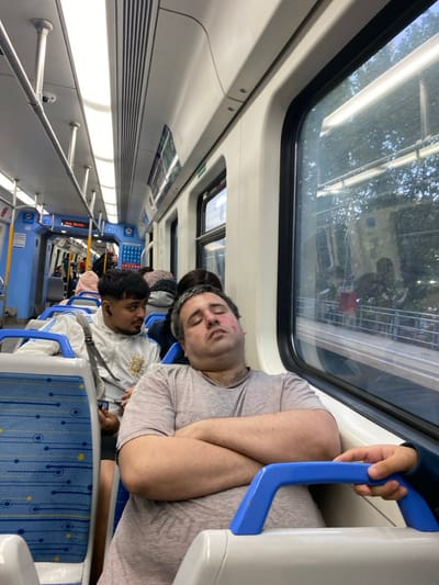 This image depicts the interior of a commuter train, specifically located within the Partido de Quilmes, Argentina. In the foreground, a middle-aged male with short, graying hair and facial redness is asleep in a light-colored train seat, leaning back with his arms crossed over his chest. He wears a light grey short-sleeved shirt. To his left and slightly behind, a younger male with dark hair and a short beard is seated, wearing a white tracksuit adorned with the Argentine national emblem. He has a black earbud in his right ear, holds a smartphone in his lap, and is observed looking directly towards the sleeping man.
Further into the train car, additional passengers are visible, some seated and some standing. The train's interior features modern blue and white patterned seats with blue grab handles, light blue wall panels, and an off-white ceiling. Overhead linear fluorescent lights illuminate the cabin, alongside air ventilation grilles. Silver vertical and horizontal handrails are present throughout. A ceiling-mounted security camera is visible in the upper left. An electronic display board shows text including "Sob. Bs.as." and "M 49." Through the large window on the right, a blurred view of an outdoor environment, possibly a station platform with trees and building structures, is visible, interspersed with reflections of the train's interior. A hand, likely belonging to another passenger, is seen gripping a blue seat handle in the lower right corner of the frame.