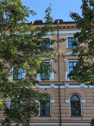 The image shows the facade of a building in Oulu, Finland, partially obscured by green trees. The building has a beige-colored exterior with white decorative elements around the windows and cornices. There are multiple windows visible, some of which are rectangular and others are arched. The windows appear to be dark-colored and some have blinds. A downspout runs vertically along the facade. The sky above is clear and blue. The overall impression is of an urban building with surrounding vegetation.