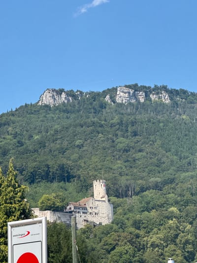 The image shows a view of the Honsteinercliff and the nearby Oensingen Castle in the Swiss municipality of Oensingen, canton of Solothurn. The castle is a medieval fortress built on a rocky outcrop. The Honsteinercliff, a prominent limestone cliff, forms the backdrop to the castle. The image captures the castle and the surrounding lush green forested hills under a clear blue sky with some wispy clouds and a faint contrail. In the foreground, a signpost with the text "oensingen" and a red circular symbol is visible. The scene is a scenic representation of the natural and historical landmarks of Oensingen.