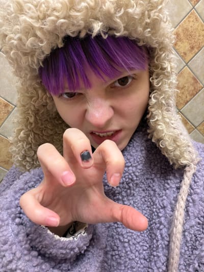 A close-up photograph, taken in the city of Lubnya, Russia, features an individual with short purple hair and brown eyes, partially obscured by a light brown, curly textured hat. The individual's face displays a snarling or growling expression, with the mouth slightly open revealing teeth. The person is wearing a purple, textured jacket. In the foreground, the individual's left hand is raised, with fingers curled into a claw-like gesture; the nail on the middle finger is dark, appearing chipped or unevenly applied. The background consists of a wall tiled with alternating light brown and beige square tiles.