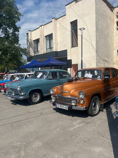 The image shows a sunny day in Skravena, Bulgaria, with a lineup of vintage cars parked on asphalt. A modern building with a beige facade and dark windows stands in the background, with a large green tree to the left. Two blue tents are set up in front of the building.

The foreground features several classic sedans. The most prominent is a shiny orange car with chrome accents and a license plate reading "CO 9835 AX". To its left is a teal-colored sedan with a similar vintage design and a license plate "CB 2121 BK". Behind it, a white car and a red car are partially visible, also appearing to be older models.

The cars are parked in a row, suggesting an event or exhibition. The ground is asphalt, with some white markings visible. The sky is blue with scattered clouds. The overall scene is one of a bright, outdoor gathering focused on classic automobiles in a semi-urban or town setting.