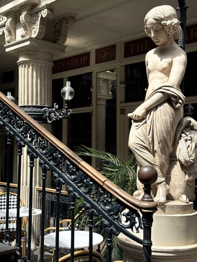 The image shows a statue of a young woman in classical attire standing next to a staircase in Nantes, France. The statue is made of stone and depicts the woman with her head bowed and her arms crossed. She is wearing a draped garment that reveals her chest and legs. The staircase is made of black metal with wooden handrails. In the background, there are shops with signs that read "THE CARD'S SHOP" and "THE AR." The scene appears to be an outdoor public space, possibly a courtyard or a plaza. The lighting suggests it is daytime, with sunlight casting shadows on the statue and staircase. There are tables and chairs visible near the staircase, indicating a possible seating area for a cafe or restaurant. A potted plant with large green leaves is also present in the scene, adding a touch of nature to the urban environment.