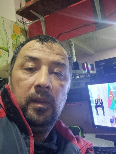 A close-up shows a man with dark hair and a beard. The man is wearing a dark jacket with red trim. He appears to be lying down, looking towards the camera. Behind him, a television displays a news program, including a flag, possibly of Azerbaijan, and a person in a suit. The background includes a red wall, a table with a patterned surface, and some electronics.