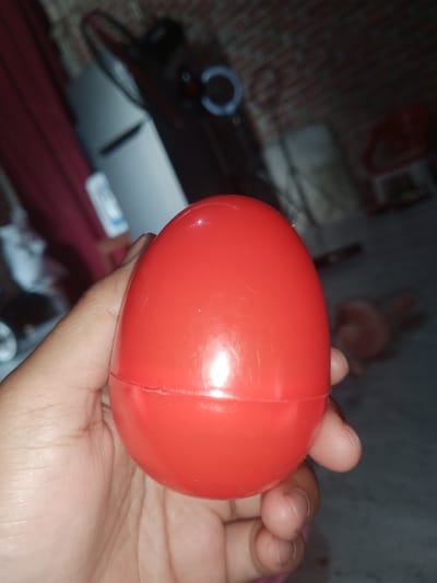 A close-up, slightly blurry, eye-level shot shows a person's left hand holding a bright red, oval-shaped plastic capsule. The capsule appears to be made of two halves that are not perfectly aligned, with a visible seam running around the circumference. The hand, with visible fingernails, is positioned to the left of the capsule, supporting it from below. The background is dark and indistinct, but suggests an indoor environment with what looks like a brick wall and furniture. The overall impression is a candid snapshot, possibly taken in a residential setting in Cotseutui, Indonesia, due to the mention of the location.