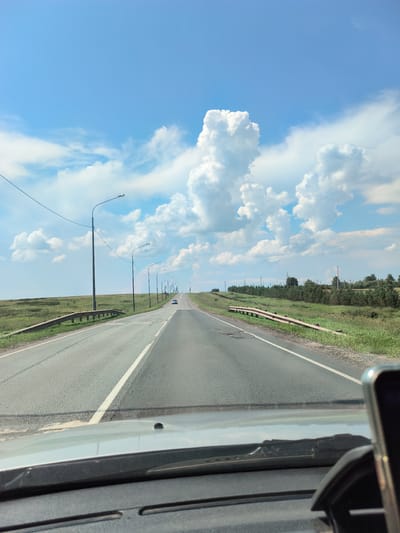 The image, taken from the perspective of a vehicle's dashboard, captures a view of a paved, two-lane road stretching into the distance. The road is flanked by grassy fields and a line of trees on the right side. Overhead, a bright blue sky is adorned with large, fluffy white clouds. Streetlights are spaced along the left side of the road. A silver car is visible in the distance, traveling on the road. The foreground shows the interior of the vehicle, including the windshield and a portion of the dashboard with a phone mount. The scene depicts a rural or highway setting, possibly near Старокутлумбетьево, Russia, given the context.