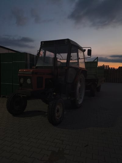 A Zetor tractor is parked on a paved surface in front of stacked green crates and wooden pallets, under a dusky sky. The tractor is red and appears to be a model from the 1970s or 1980s, with a visible cab and a trailer attached. The scene is set at twilight, with the sky displaying hues of blue, grey, and orange, indicating a sunset. The environment suggests an industrial or agricultural setting, possibly a storage yard or depot. The image composition focuses on the tractor and its immediate surroundings, with the background elements of crates and pallets providing context. The lighting is dim, with shadows cast on the ground, further emphasizing the time of day.