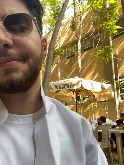 The image depicts an outdoor scene in قرية الضبية, Syria, captured under bright sunlight, suggesting daytime. In the immediate foreground, the upper left quadrant features a male individual partially visible from the chest up. He wears dark-lensed, gold-framed sunglasses, has a dark beard and mustache, and is dressed in a white crew-neck t-shirt beneath a white collared jacket or garment. His gaze is directed slightly to the left, and his mouth is closed.
The midground reveals an outdoor cafe or seating area. A large, weathered beige umbrella, bearing the black 'NESCAFE' logo, is centrally positioned and partially unfurled, showing some damage or wear. Several slender trees with green foliage are scattered throughout the scene, casting prominent shadows across the background building. White plastic chairs and light-colored tables are visible, occupied by at least three other individuals. One person is seen from behind, leaning forward at a table, wearing a light-colored top. Another individual with a dark beard and dark cap is visible further right, seated at a table. Small glasses or cups are present on the tables.
The background consists of a multi-story building with a light brown or beige facade. The building features multiple evenly spaced windows with dark frames, reflecting the bright outdoor light and displaying shadows cast by the trees.