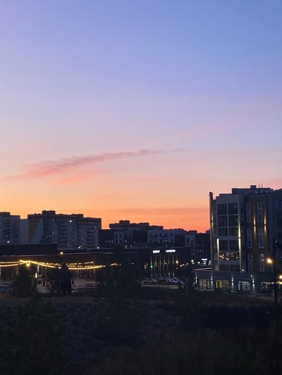 The image captures an urban or suburban scene at twilight, likely dusk. The sky occupies the upper portion, displaying a gradient of colors from a soft purple-blue on the left to warm orange and pink hues on the right, typical of sunset. A long, faint, wispy cloud stretches horizontally across the colorful expanse.

Below the sky, a skyline of multi-story residential or mixed-use buildings forms a prominent dark silhouette against the vibrant light. Many windows within these buildings are illuminated, casting warm glows. In the lower right foreground, a dark expanse of grass or foliage is visible. The scene is lit by various artificial lights, including individual lampposts and decorative string lights that extend in a line, suggesting a pathway or public promenade.

Close inspection of cropped sections reveals at least one, and possibly two, human figures as dark silhouettes walking along this illuminated path, suggesting an evening stroll. The overall atmosphere is peaceful and serene, capturing the transition from day to night in a populated area. No discernible text is visible on any objects or buildings. This view is consistent with a modern residential area in Chemoshur, Russia.