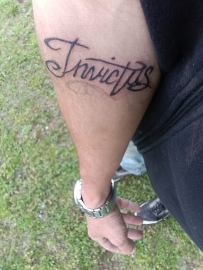 The image shows a person's arm with a tattoo that reads "Invictus" in a cursive font. The tattoo is located on the inner forearm. The person is wearing a metal watch on their wrist. The background is green grass, and parts of a dark-colored shirt, blue jeans, and a dark shoe with white trim are visible. The location is outdoors and appears to be a grassy area. There is no indication of the location being within the city of Stilwell, United States.