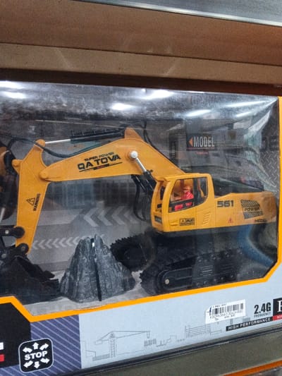 A toy excavator in its packaging features a bright yellow "QA TOVA" boom with a warning sticker, a gray rock-like accessory, and black treads. The cab includes a driver and the number "561". Packaging details include "SUPER POWER," "MODEL," and a "STOP" icon. A sticker indicates a 2.4G frequency for high performance. The lower packaging also features a simple drawing of a small building.