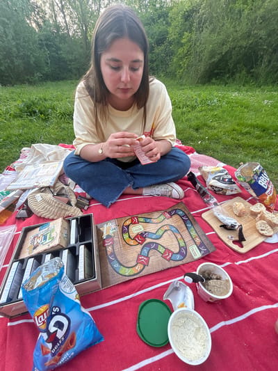 A woman sits cross-legged on a red and white striped blanket in a grassy area, possibly Marquette-lez-Lille, France, engaging with a game of "Time's Up". She holds cards in her hands. A board game, the game box, and card storage are visible on the blanket. Various picnic items such as potato chips, sliced bread, cheese, and pate are scattered around. A light yellow shirt and blue jeans adorn the woman. Trees and grass comprise the background.