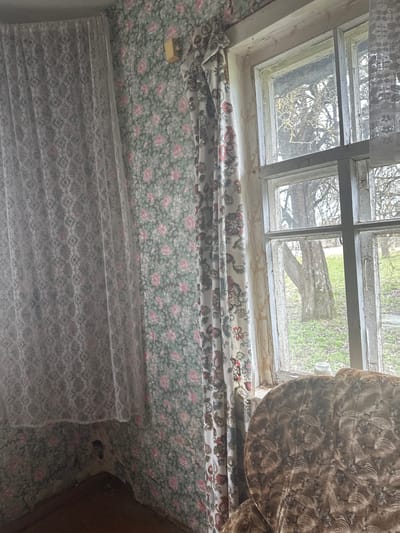 The image presents an interior scene, likely a room in a house. A window with a weathered frame is visible at the top, offering a view of a tree and foliage outside, suggesting a rural or suburban setting. The window is partially obscured by floral-patterned curtains or drapes. Below, floral wallpaper covers the wall, and a lace curtain is draped further down. Part of a piece of brown patterned furniture is seen on the right side of the frame. The overall impression is one of age and disrepair. There are no people visible in the image. The location is in the Студзёнкаўскі сельскі Савет area of Belarus, per the prompt.