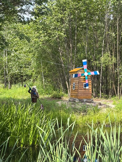 The image depicts a wooden windmill with colorful vanes and a small window, situated on a stone base in a grassy area. To the left, a scarecrow-like figure is visible, adorned with a leopard-print outfit and a blonde wig, holding fishing rods. The scene is surrounded by lush green trees and vegetation, with a body of water in the foreground filled with reeds, reflecting the windmill and the sky. The overall setting appears to be an outdoor decorative exhibit in a park or garden, potentially in Kul'dur, Russia, given the context provided. The lighting suggests it is daytime, likely under sunny conditions.