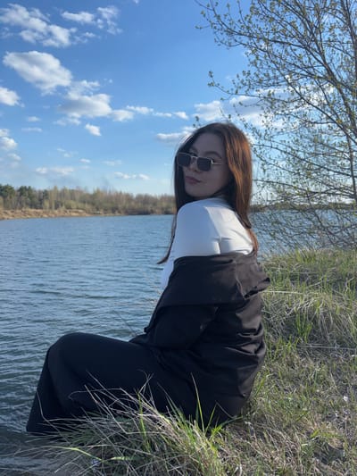A woman is kneeling near a body of water, possibly a lake, in an outdoor setting. She is wearing sunglasses, a white shirt, and a black jacket with matching pants. The environment consists of grass and foliage, with trees visible in the background. The sky above contains blue patches with scattered clouds. Given the city specified is Zaosinovo, Russia, it is likely that the picture was taken there.