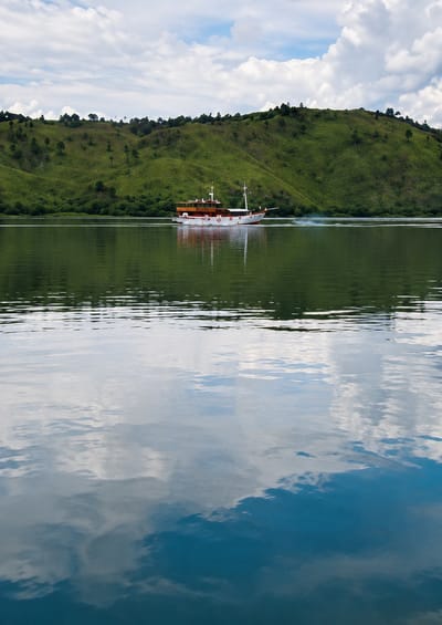 A long, narrow boat with a white hull and a brown wooden deck is traveling across a calm, green body of water. The boat has two masts and several life preservers visible along its side. In the background, rolling green hills are covered in lush vegetation and scattered trees. The sky above is partly cloudy with dramatic white clouds against a blue sky. The reflection of the boat, the hills, and the sky is clearly visible on the surface of the water. This scene is located in Turpuk Sihotang, Indonesia, likely on Lake Toba, a large volcanic lake.