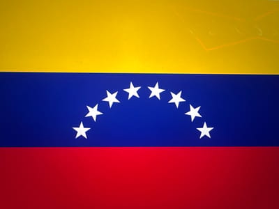 The flag of Venezuela is a tricolor of yellow, blue, and red horizontal stripes. Eight white, five-pointed stars are arranged in an arc in the center blue stripe. The upper right corner of the yellow stripe has a visible scuff mark.