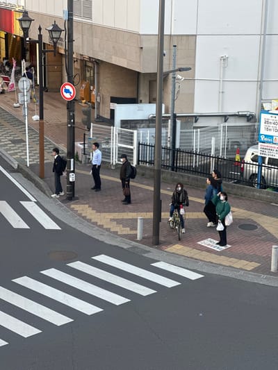 This image depicts an urban street scene in Tokorozawa, Japan, viewed from a slightly elevated perspective. The foreground features a dark asphalt road with a prominent white zebra crossing (crosswalk) and a circular manhole cover. Paved sidewalks, constructed from light brown and reddish-brown interlocking pavers, run parallel to the road on both sides.

On the left sidewalk, three males are standing. From left to right: a young male, possibly a student, wearing a dark uniform, glasses, and a backpack, facing right; an adult male in a light blue long-sleeved shirt and dark pants, facing right; and an adult male in a dark jacket, dark baseball cap, and white face mask, carrying a dark backpack and a brown bag in his left hand, also facing right. This sidewalk is lined with multiple black utility poles carrying numerous overhead cables, along with two ornate dark streetlights. A red circular 'No Trucks' traffic sign is affixed to one utility pole. Buildings with light-colored facades, some displaying internal yellow lighting from storefronts, are visible further along the street.

On the right sidewalk, four individuals are present. A female in a dark jacket and blue jeans, wearing a white face mask, is seated on a light-framed folding bicycle, facing left towards the crosswalk. Immediately behind her, two females stand close together, both with dark hair, one in a dark blue jacket and the other in a dark jacket, both facing left. Further right, a female in a light green long-sleeved top, dark pants, and a white face mask, holding a white shopping bag in her left hand, stands on a white square pavement sticker with Japanese text, looking upwards and to the right. This sidewalk is bordered by metal fences (white and black sections) separating it from a parking area, where a silver van is partially visible behind the black fence. A multi-story building with a beige lower facade and white upper facade, featuring exterior industrial pipes and vents, fronts this side of the street. A blue parking lot sign, detailing hourly rates and displaying "2024年3月2日" (March 2, 2024), is visible near the parking area. Small white cylindrical bollards are interspersed along the sidewalk edge.

The overall lighting is soft and diffused, suggesting an overcast daytime condition or late afternoon, with minimal harsh shadows.
