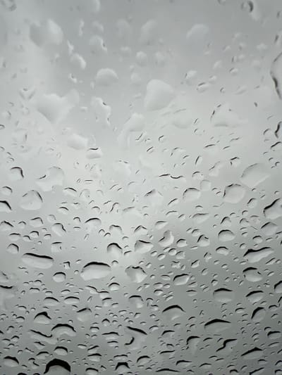 The image is a close-up, high-angle view of raindrops on a surface, likely a window, against a uniformly gray, overcast sky. The raindrops are of varying sizes, from small droplets to larger, irregular blobs, some appearing to have coalesced. The surface itself is smooth and shows subtle variations in light and shadow, which highlights the three-dimensional form of the water. There are no people, identifiable objects, or structures visible in the frame. The scene is devoid of any contextual clues to indicate a specific location, including the city of Jõhvi, Estonia. The overall impression is one of a wet, desolate, and monotonous atmospheric condition.