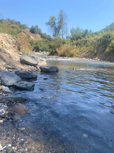 The image shows a natural landscape with a clear blue sky and a river flowing through a valley. The foreground is dominated by a rocky riverbank with several large, smooth stones partially submerged in the water. The river itself is shallow and appears to be flowing over a bed of smaller pebbles and rocks. The water surface is rippled, reflecting the sunlight and the surrounding greenery.
The background consists of steep, grassy hillsides covered in vegetation, including trees and bushes. In the distance, along the riverbank, there are some small structures that appear to be houses or sheds, with a few vehicles parked nearby. A red flag is visible on top of one of the hills. The overall impression is one of a peaceful, rural setting in a mountainous region. The location is identified as Kara-Alma, Kyrgyzstan.