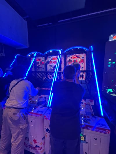 This image captures an indoor scene within an entertainment establishment in Saint-André-lez-Lille, France, characterized by predominantly blue neon lighting. Three individuals are visible, interacting with a row of "Sonic All-Stars" basketball arcade machines.
The environment is dimly lit, with dark walls and ceiling, illuminated primarily by the vibrant blue LED strips outlining the frames of the arcade games. On the far right, a vertical arcade cabinet displaying a "Space Invaders"-style game with pixelated aliens is partially visible.
In the foreground, a person wearing a dark t-shirt and dark pants is positioned at the middle basketball machine. This individual holds a small basketball with both hands, aimed towards the hoop, indicating an active play session. To their left, another person, wearing a white long-sleeved shirt and light-colored cargo pants with visible pockets, stands at the leftmost basketball machine, appearing to lean forward over its console. A dark strap, possibly from a bag, crosses their back. The console for each machine features "SEGA AMUSEMENTS" branding and "SONIC ALL-STARS" graphics. Digital scoreboards on each machine display "SCORE," "TIME," and "HIGHEST" in red numerals, with one displaying "59" for score and "33" for time. A third person is partially visible on the far left, their left shoulder and arm in dark clothing. The machines are designed with a backboard, a hoop, and a ball retrieval system.