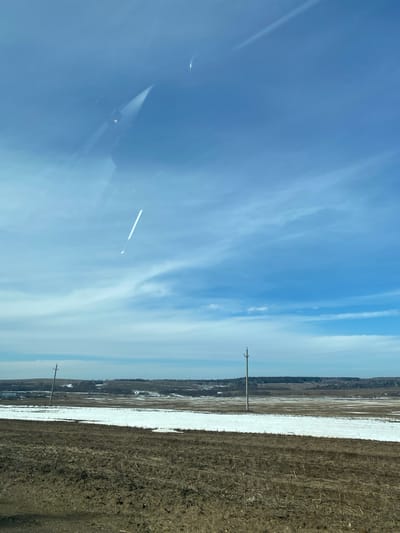 The image depicts a rural landscape on a sunny day, likely in the vicinity of Буль-Кайпаново, Russia given the prompt. A clear blue sky with scattered clouds occupies the left portion of the image. The right side of the frame shows a field, with areas of snow cover interspersed between patches of brown soil. Two utility poles are visible, sparsely spaced along the border between the field and the sky. Some trees are visible near the horizon line. Reflections are seen in the glass the photograph was taken through, adding artifacts.