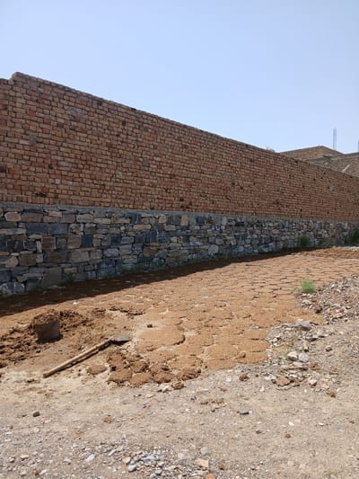 The image depicts an outdoor construction scene in Zhob, Pakistan. A tall, red brick wall dominates the background, with a lower section of stone construction below it. The ground in the foreground is covered with a mixture of soil, gravel, and what appears to be dried material, possibly manure, spread in patches. A metal bucket filled with a brown substance and a shovel are situated on the ground. The sky is clear and blue. The overall impression is one of ongoing construction or land preparation in a dry, arid environment.