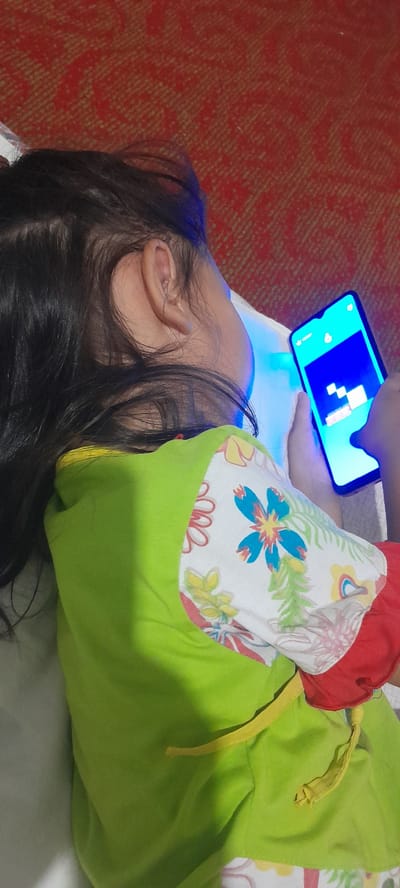 A young child lies on their side, engrossed in a mobile phone. The child's head is to the left, with dark hair obscuring their face, though one ear is clearly visible. They are wearing a bright green hooded jacket with a colorful floral pattern on the shoulders, featuring red, blue, yellow, and green flowers against a white background. A yellow drawstring hangs from the jacket. The child is holding the phone, which displays a game with blue and white geometric shapes. The blue light from the phone casts a glow on the child's face and the white surface they are lying on, likely a bed. The background features a textured red surface, possibly a rug or carpet. The scene appears to be indoors, given the artificial light from the phone. The child's posture and focus suggest deep engagement with the game. There is no discernible text in the image, and no information about the time of day or weather is available. The setting appears to be a comfortable, possibly domestic, environment. The child's emotion is one of concentration.